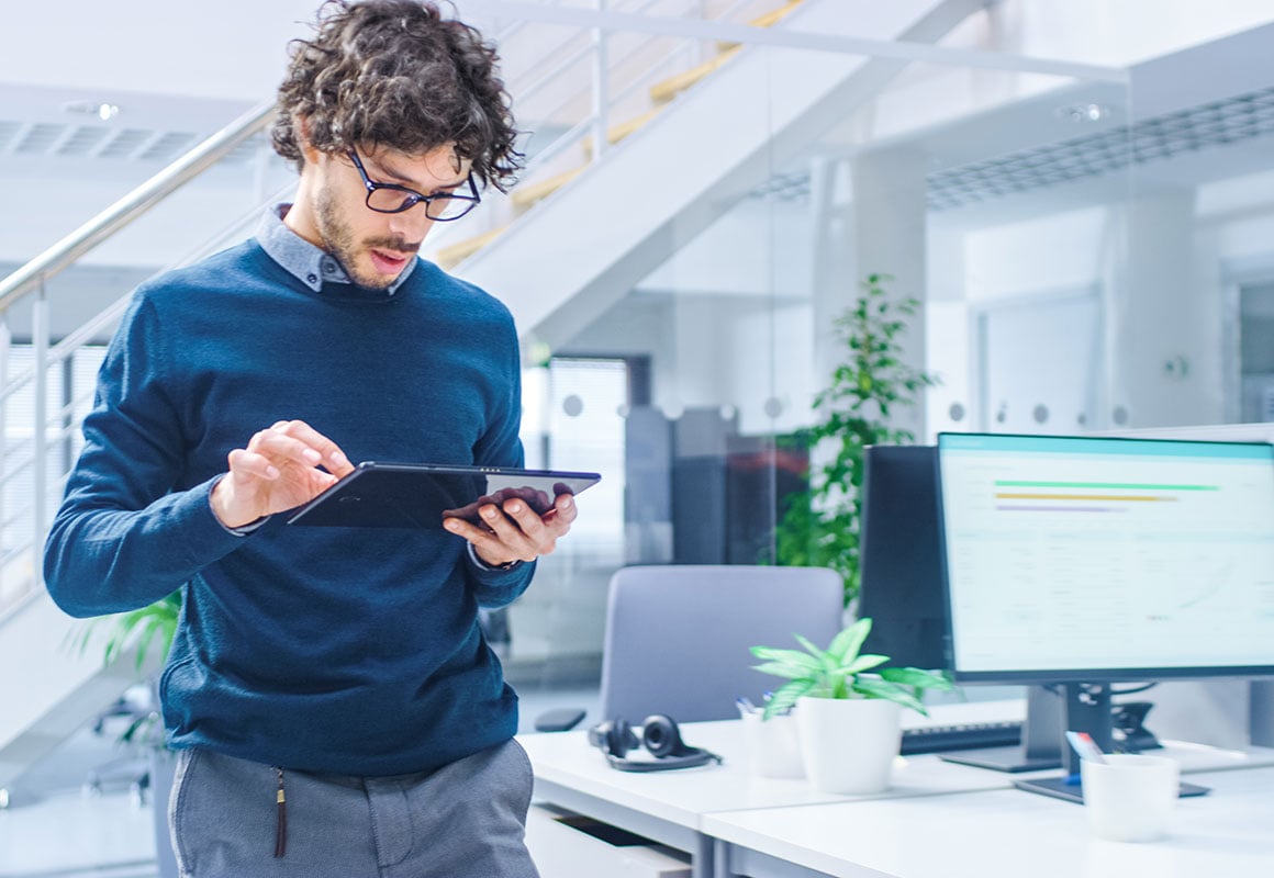 Ein Mann steht auf der linken Seite des Fotos, er hält ein Tablet in der Hand und sieht da drauf, er steht in einem hellen, modernen Büro