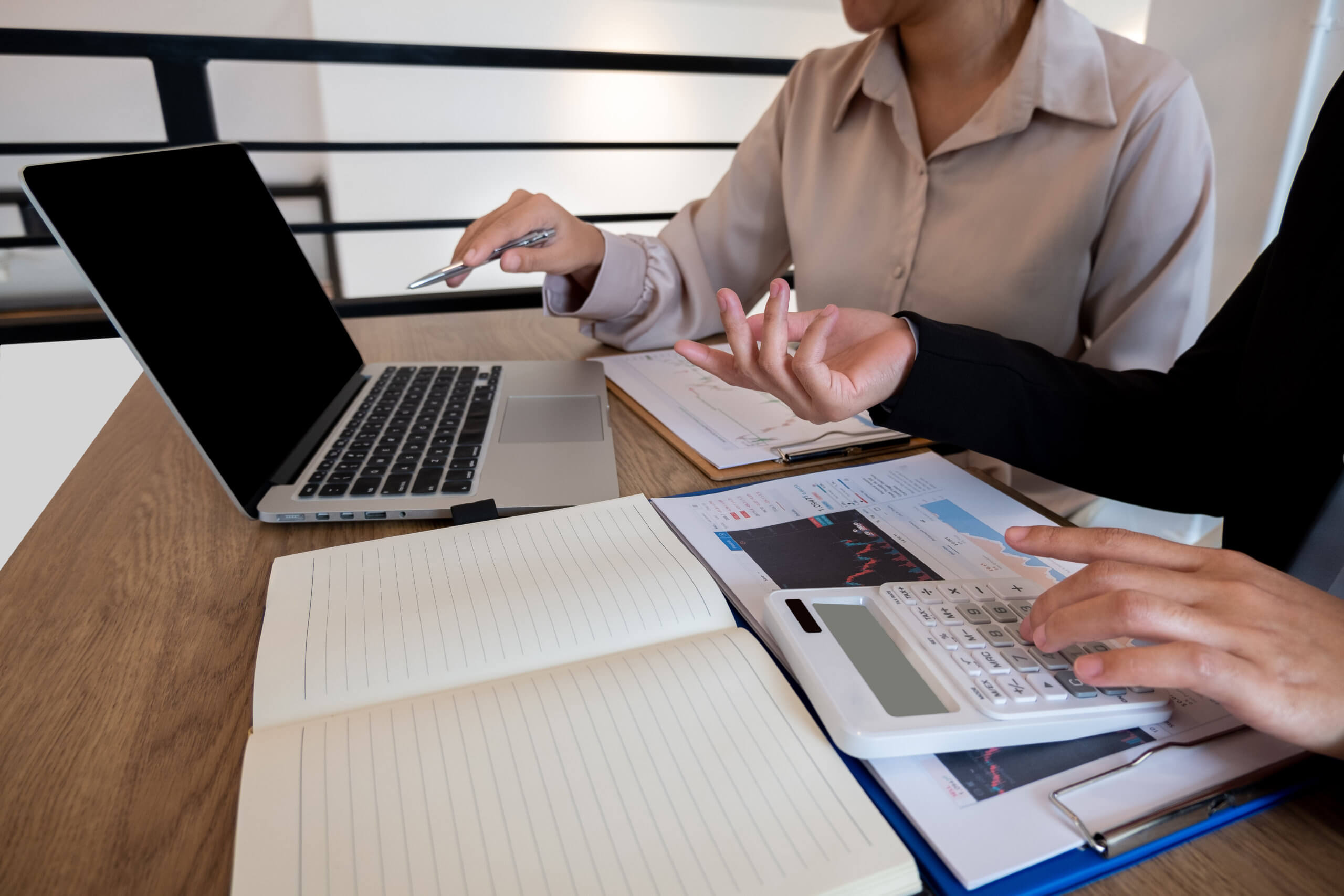 Schreibtisch mit Laptop, Dokumenten und Taschenrechner sowie zwei Personen, die sich am Schreibtisch über die Vergütungsstudie unterhalten.