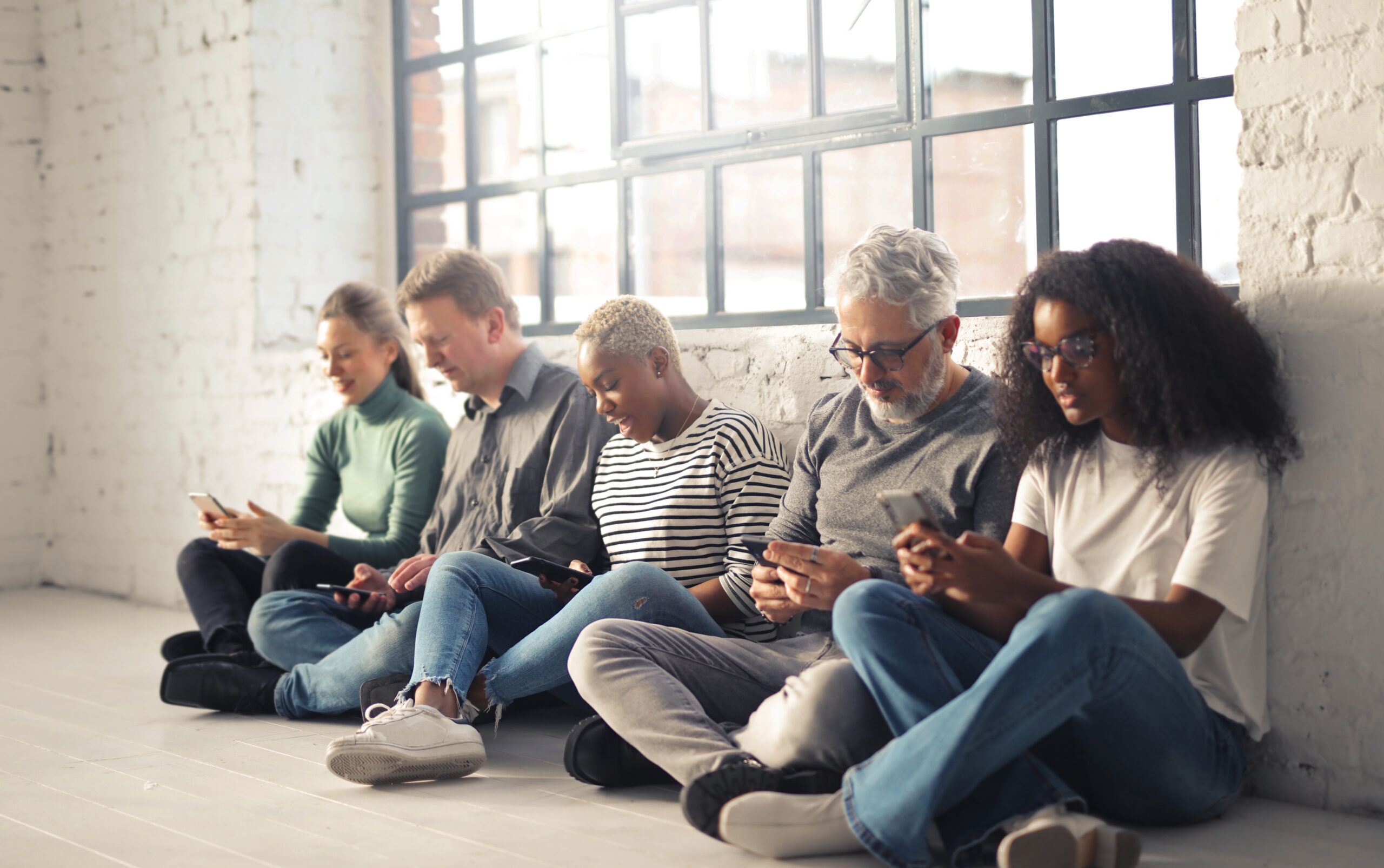 Ein Gruppe junger und älterer Mitarbeitender sitzen auf dem Boden und schauen auf ihre Handy. Sie sind ein Sinnbild für Generationenmanagement. 