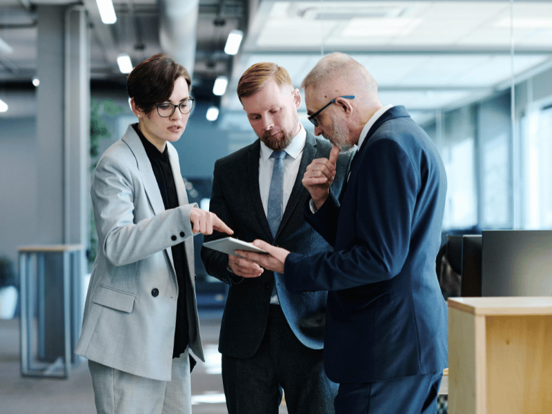 Eine Frau und zwei Männer stehen im Halbkreis und gucken auf ein Tablet.