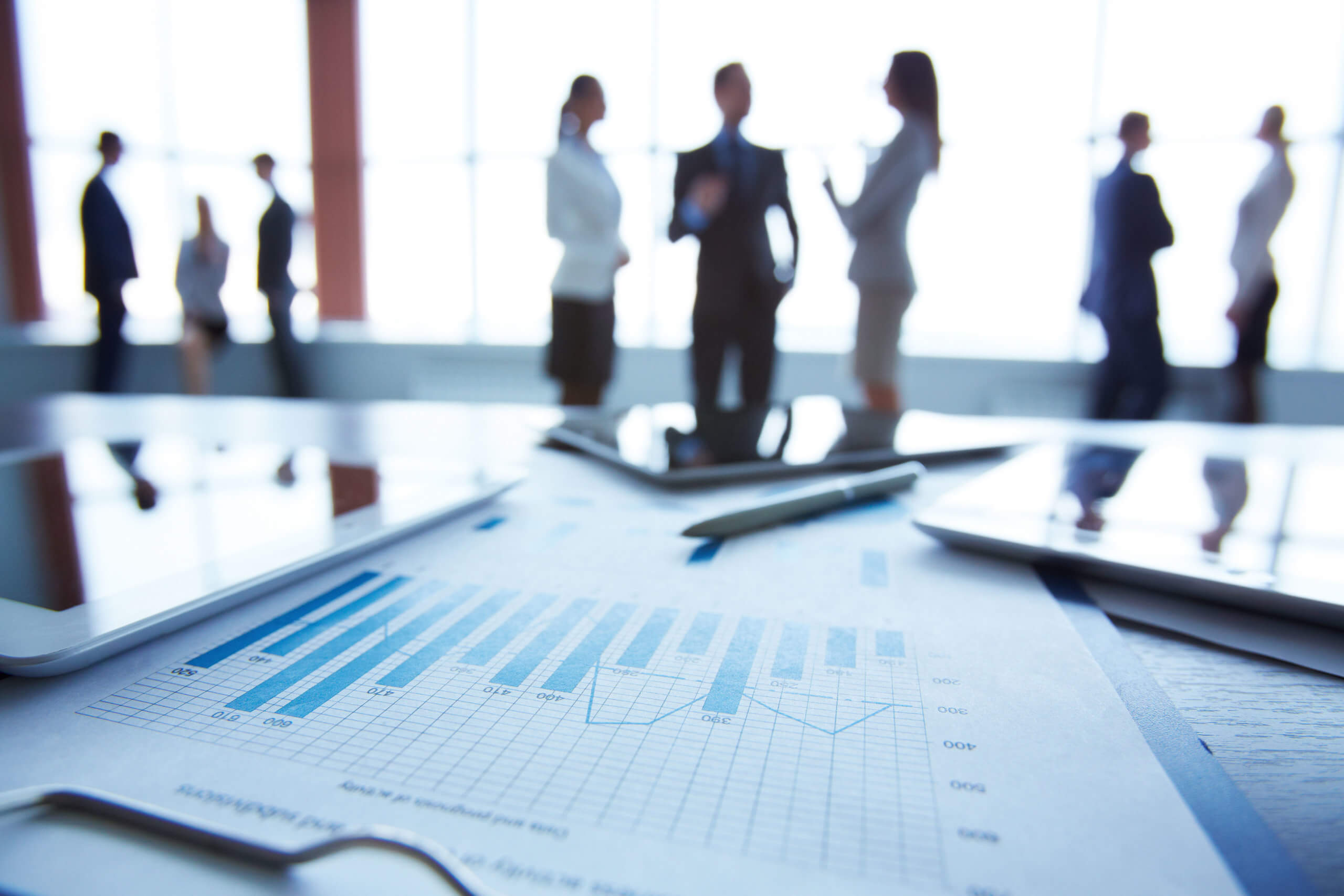 Im Vordergrund liegen Dokumente und Tablets und im Hintergrund sieht man, wie sich Leute im Raum bewegen und unterhalten.