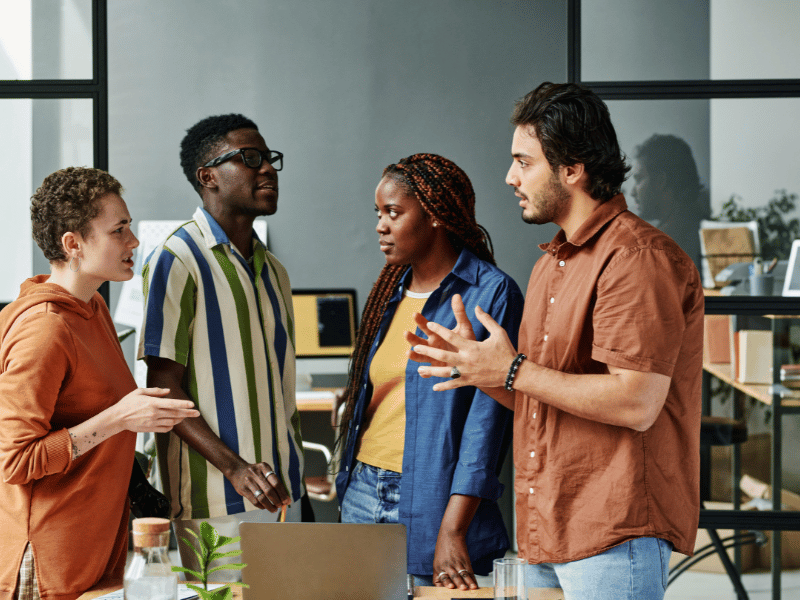 Vier Kollegen stehen an einem Tisch im Büro und diskutieren miteinander. Ein Symbolbild für eine Szene aus einem Workshop mit der Walt Disney Methode.