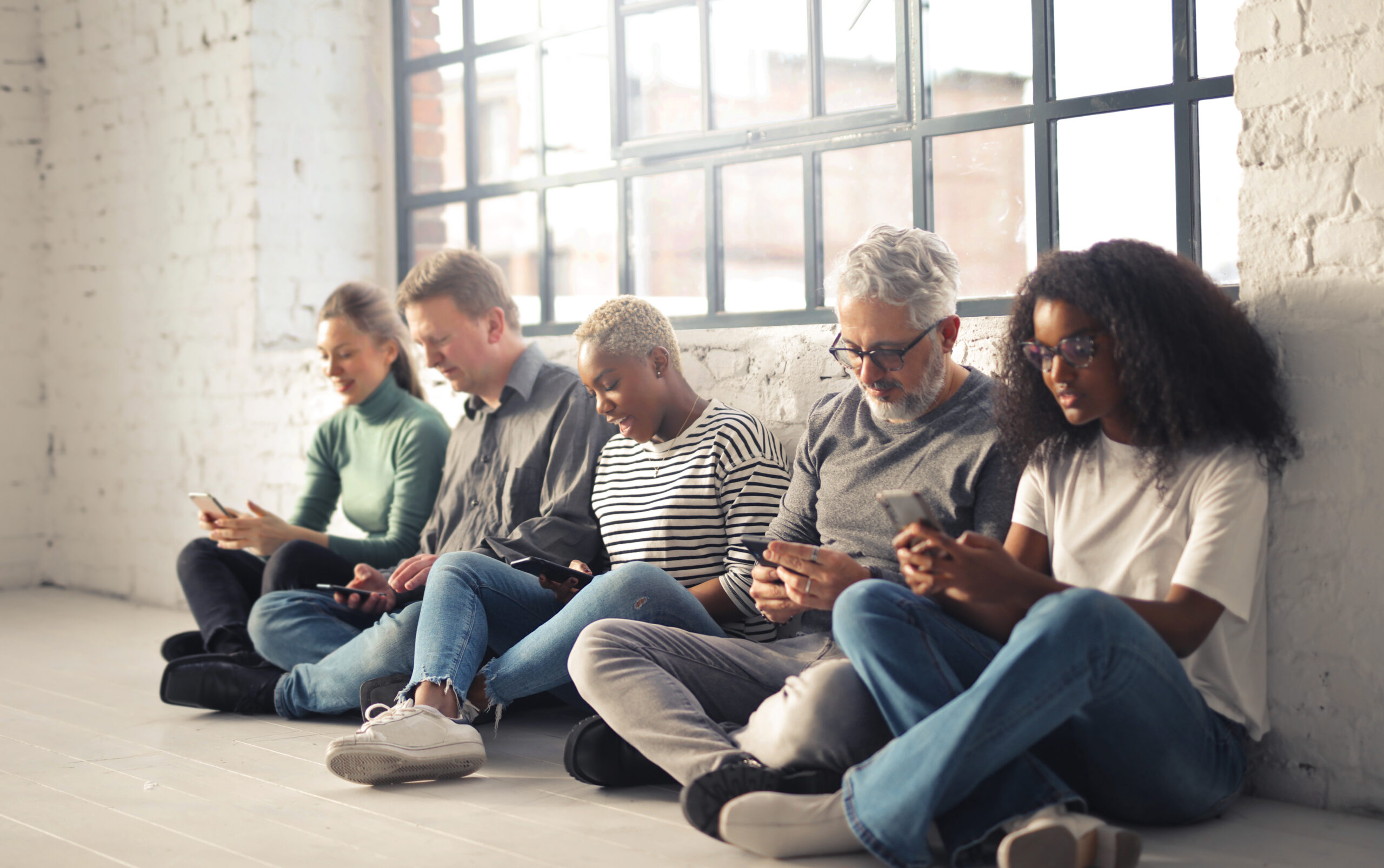 Eine Gruppe von Arbeitskolleg*innen verschiedener Altersgruppen sitzen vor einem Fenster auf dem Boden und schauen auf ihre Smartphones.