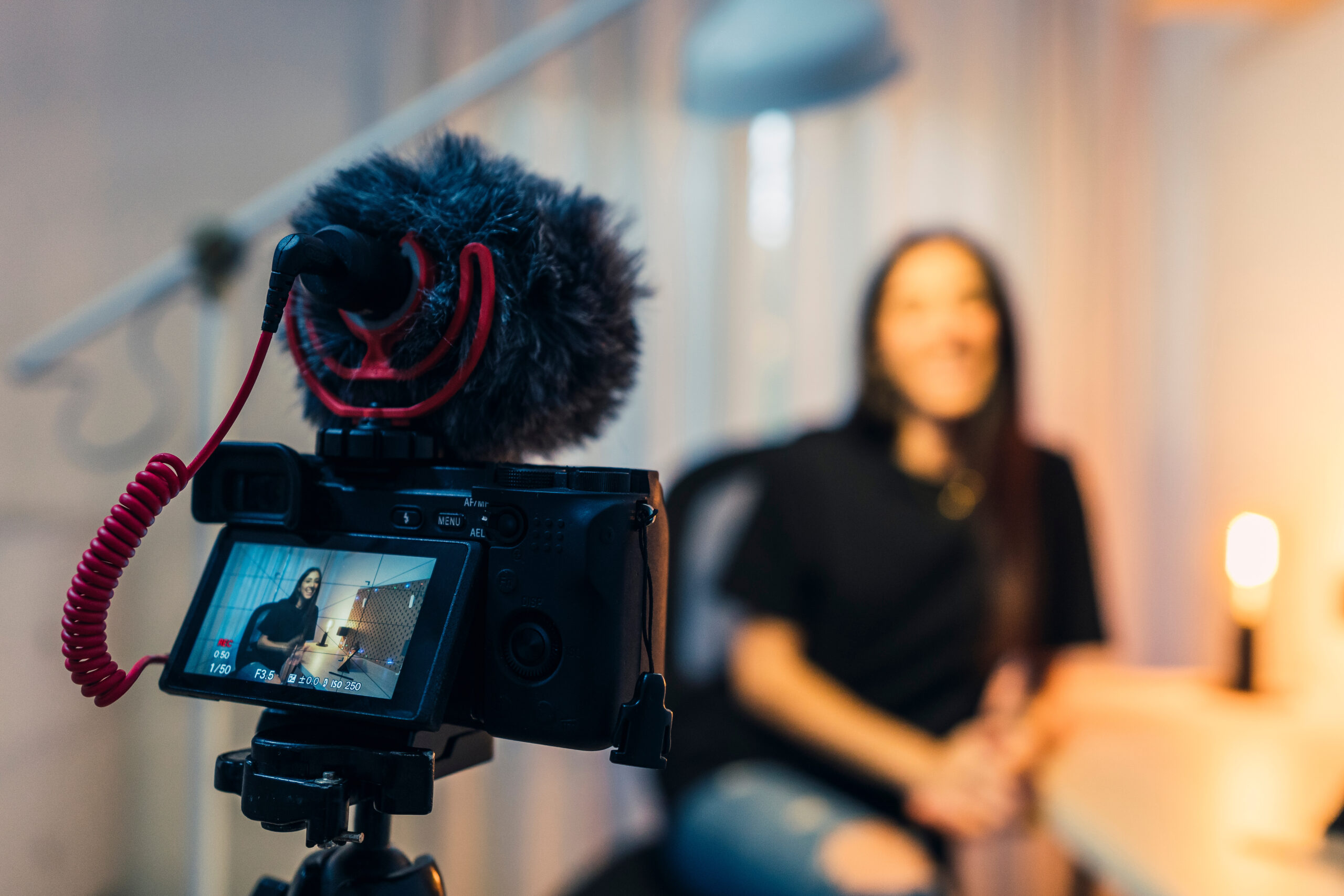 Im Vordergrund sieht man eine Kamera mit einem Mikrofon und im Hintergrund eine Frau, die interviewt wird.